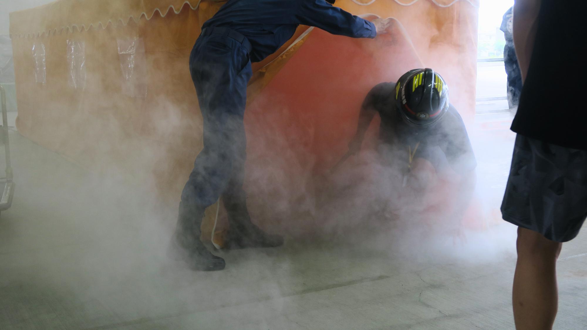 参加者がヘルメットをかぶり、訓練用の白煙が充満しているテント内から訓練人形を救出している写真