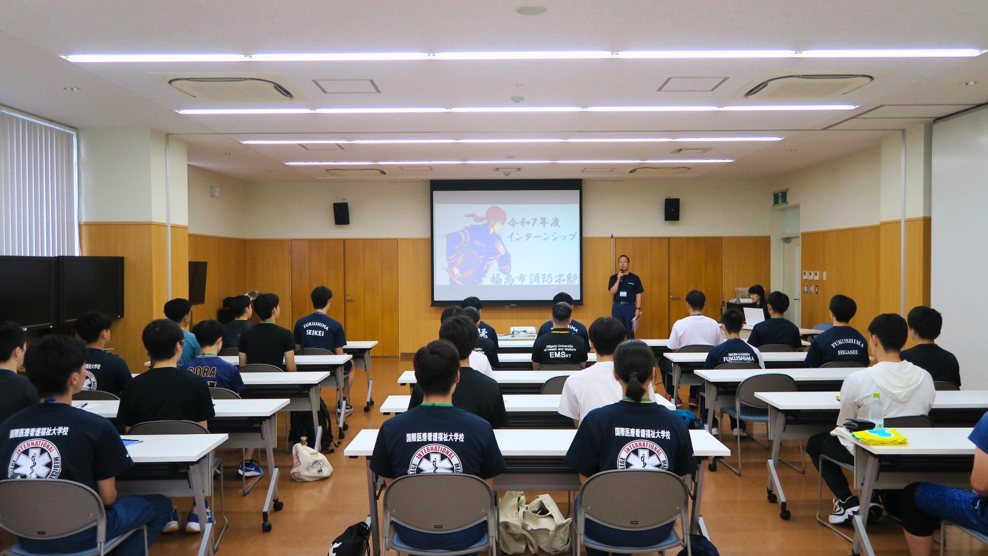 会議室内でマイクを持って説明をする消防職員と、説明を自席で聞く参加者の写真