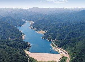 深緑の山々が連なるV字谷の地形に沿って、青い水を湛えた細長い人造湖が広がり、手前の大きなダムと湖岸を走る道路の様子を捉えた空撮写真