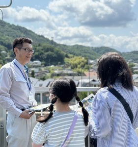 周囲の山並みと街並みを背景に白い作業服姿の男性が、参加者に笑顔で説明をしている様子の写真