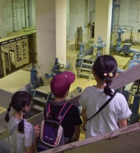 参加した子どもたちが大きな建物内の配管やバルブ、制御盤などの機械設備が並ぶ地下室のような場所を、階段の上から見学している様子の写真