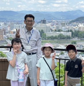 山々に囲まれた市街地の高層ビル群を背景に、ピースサインをする作業服姿の男性一人と三人の子どもたちが笑顔で写っている写真