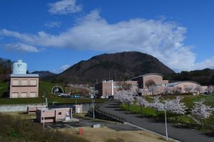 大きな山を背景にした敷地内に、手前左に円筒状のタンクを持つ四角い建物、中央奥に茶色い屋根と丸みを帯びた構造物を持つすりかみ浄水場の写真