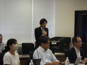 スーツ姿の男性がマイクを片手に立って話をしている様子の写真