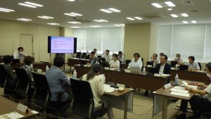 会議室で、長テーブルを囲んで十数名の参加者が着席しており、奥のスクリーンに文字のスライドが映し出され、手前の参加者が熱心に資料を見ている様子の写真