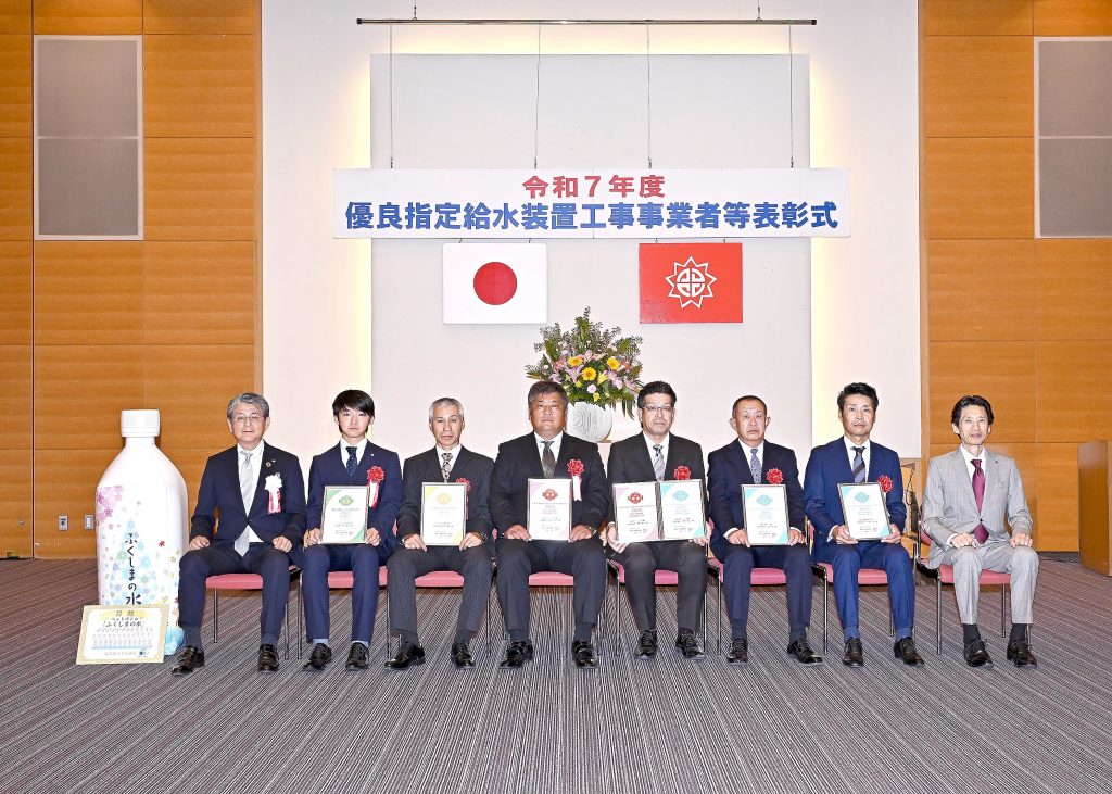 「令和7年度優良指定給水装置工事事業者等表彰式」と書かれた横断幕の前で、中央に国旗と市の旗、その下に花束が飾られた会場にて、スーツ姿の8人の男性が着席し、表彰状を手にしている集合写真