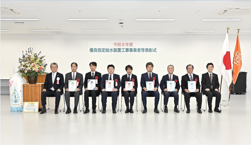 「令和8年度優良指定給水装置工事事業者等表彰式」と書かれた横断幕の前で、その下に国旗と市の旗、花束が飾られた会場にて、スーツ姿の9人の男性が着席し、表彰状を手にしている集合写真
