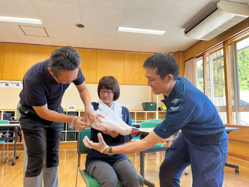 椅子に座った方の右手に三角巾を首から掛け三角巾を付ける練習をしている方と説明をしている講師の実習の様子の写真