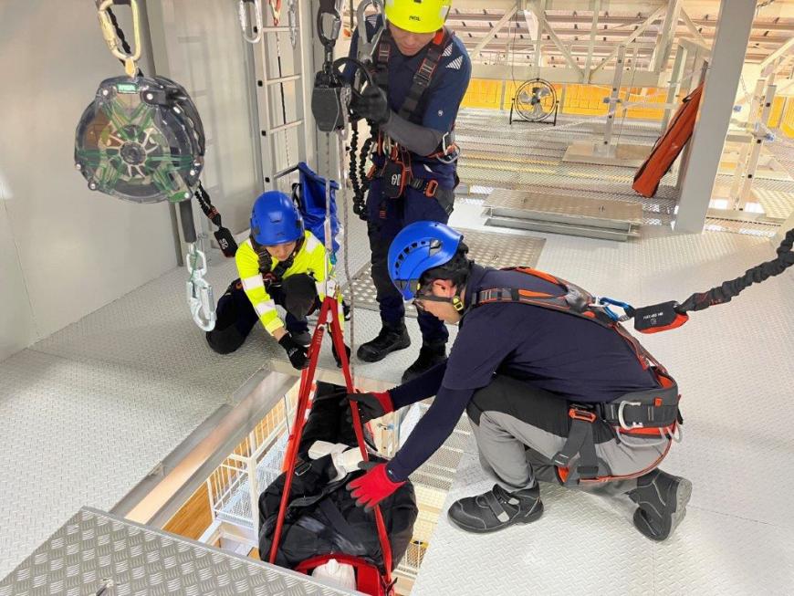 大型訓練設備の上からベルトで吊り下げられた荷物を道具で下げようとしている実習の写真