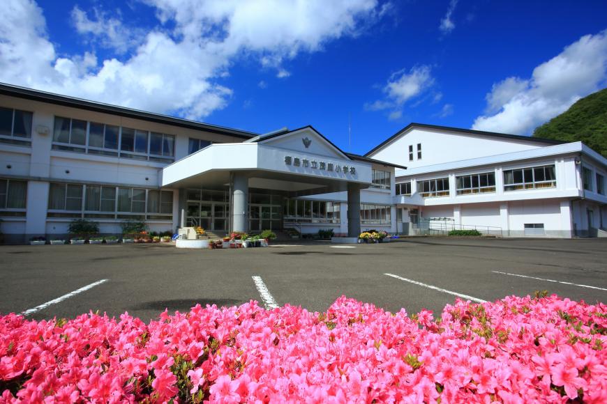 前方にピンク色のつつじの花が咲き白を基調とした2階建ての建物と右側には白の外観の体育館のある旧茂庭小学校の写真