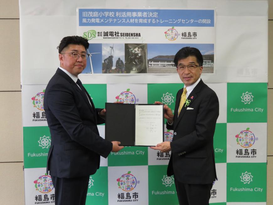 福島市のバックパネルの前で紺色のバインダーに挟まれた書類を2人で持ち記念撮影をしている市長と関係者の男性の写真