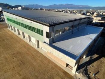 手前の窓側に緑色のネットが設置された完成した体育館を、山並みや住宅街が見える高台から撮影した写真
