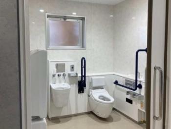 白色で統一された奥に喚起窓が設置され、壁に手すりが設けられた洋式トイレと手洗い場が設置されたバリアフリートイレの写真