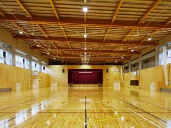 奥にステージがあり左右にバスケットゴールが設置され、天井に設置された照明が床に反射している体育館内の写真