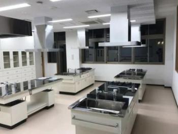 奥に窓ガラスがあり、左側の壁に食器棚、中央に4台の作業台が設置された調理実習室の写真