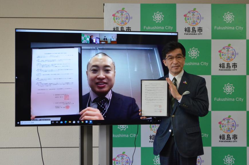 市長とモニターに映っている株式会社バカン(VACAN)の関係者の男性が、協定書を持って写っている記念写真