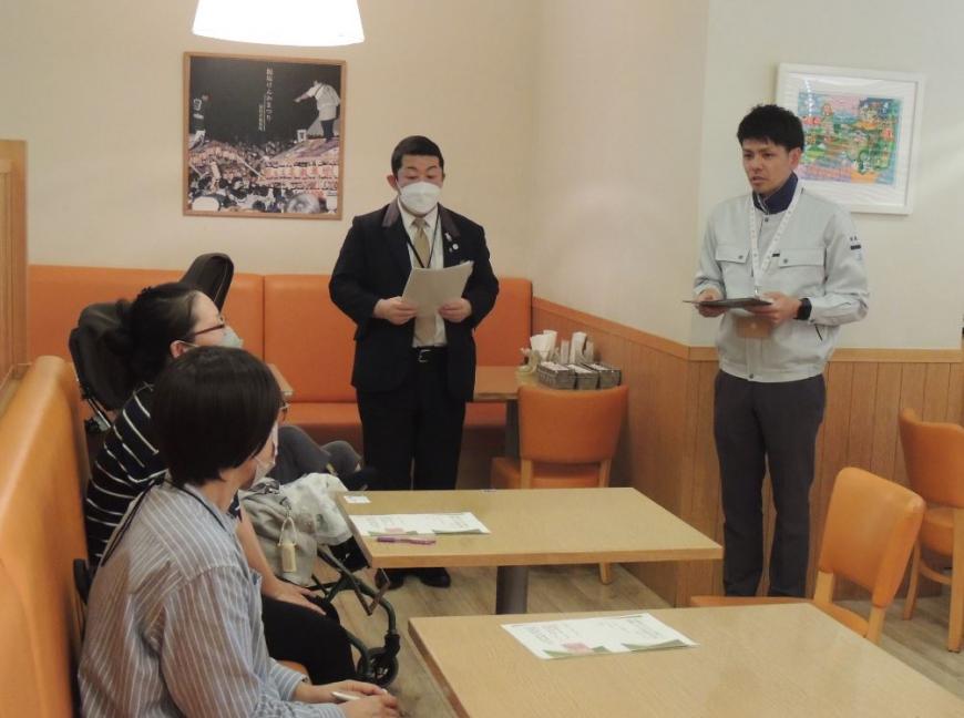テーブルが二つ並び、参加者達が椅子に座っている前方に、男性2人が参加者達の方を向き、手元に資料を持って立っている写真