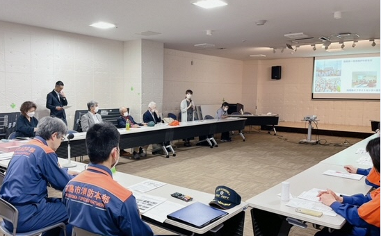 前方のスクリーンに資料が映され、左側の自席に立った女性がマイクを持って話をしているミーティングの様子の写真