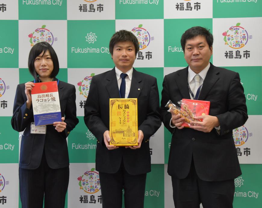 福島市のバックボードの前で3名の職員の方が長崎のお菓子のお土産をそれぞれ持って写っている写真