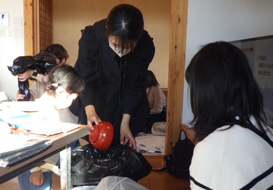 黒いごみ袋の中にお椀に入ったものを入れており、幼い女の子と参加者のママがトイレ実験をしている様子の写真