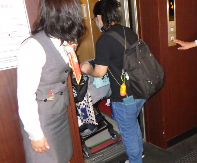 関係者の女性がエレベーターのドアを開け、ベビーカーを押している母親が、エレベーターの中に入っている写真