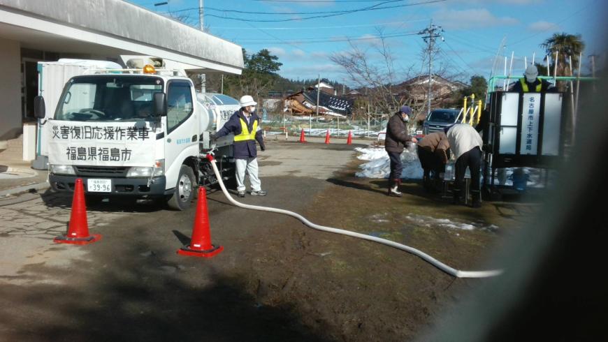 降り積もった雪が敷地の角に残っている場所で、災害復旧応援作業車福島県福島市と書かれた横断幕がつけられた給水車からホースが伸び、給水支援をしている様子の写真