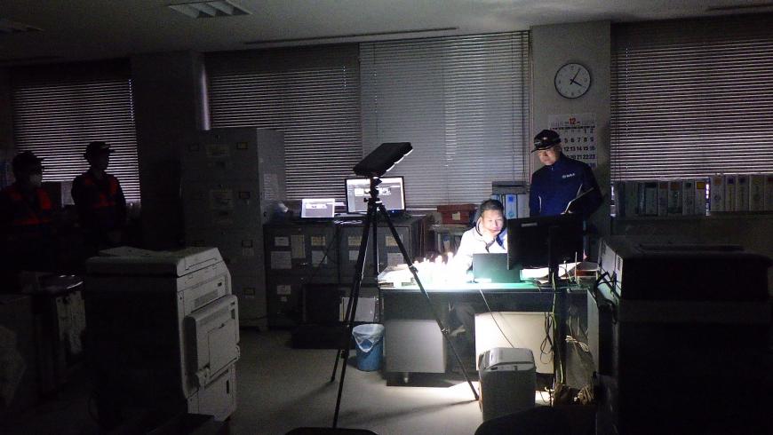 暗闇の庁舎内で一つのライトのみ照らされ、職員の男性一人は机の前に座ってパソコンを見ながら作業し、側に資料を持って男性がパソコンの画面を見ている北信支所の訓練の様子の写真