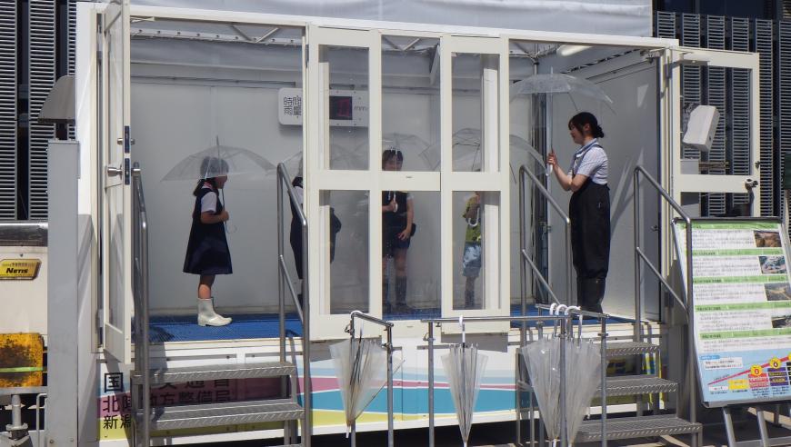 降雨体験装置内で児童数名が傘を差し、豪雨体験をしている様子の写真
