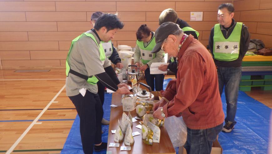 長机の上にパウチに入った非常食や、やかん、ポットが置かれており、「食事係」の緑のビブスをつけた関係者の方々が、非常食の準備を行っている様子を写した写真