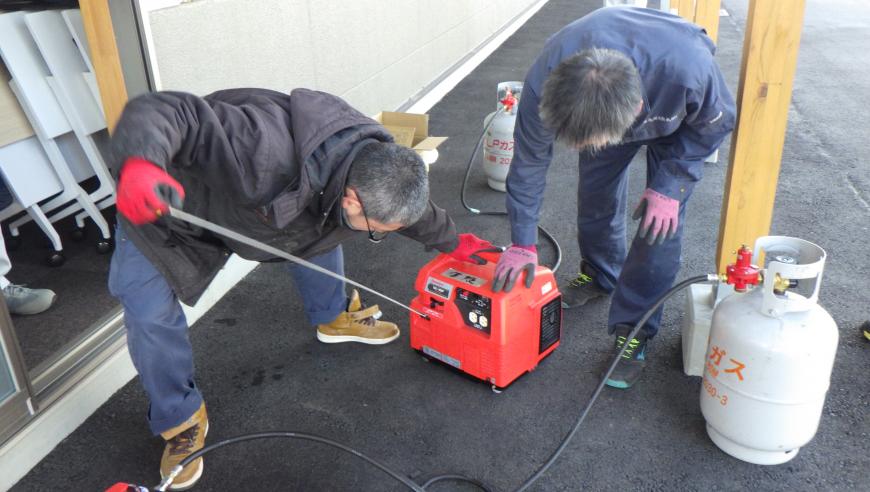 ガスボンベが置かれその横で関係者の男性2名でポータブル発電機を使う準備をしている様子の写真