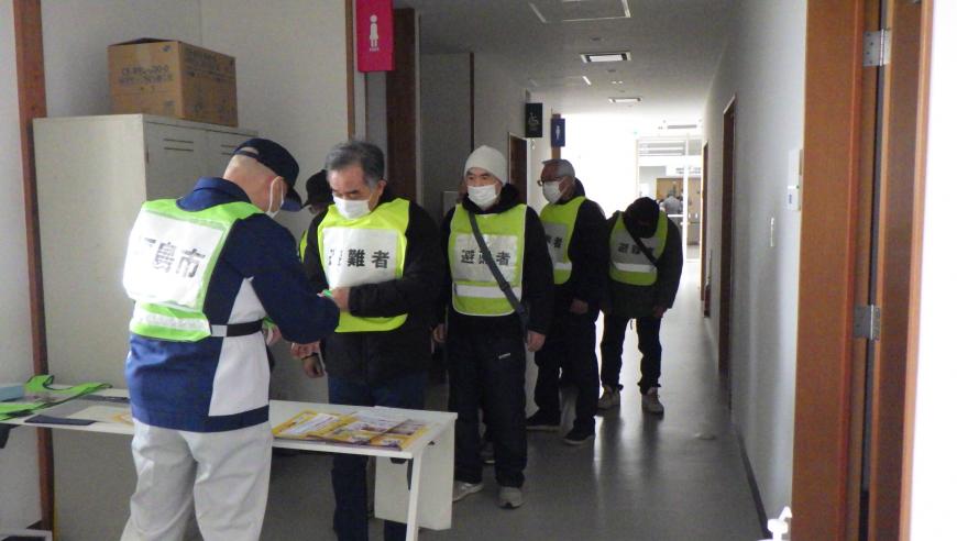 避難者のゼッケンをつけた関係が並んでおり、福島市のゼッケンをつけた関係の男性が受付をしている様子の写真