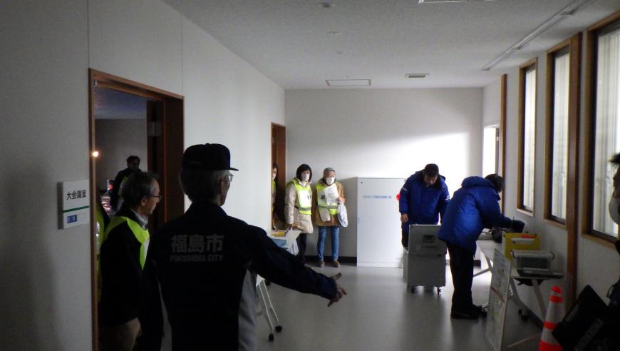 廊下の壁側に長机が設置され、関係者の方々が機械を運び準備をしている様子の写真