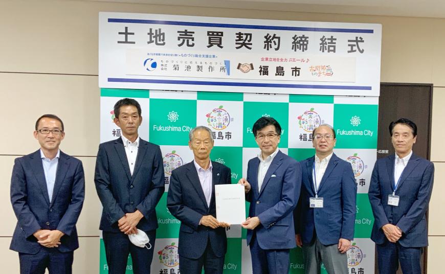 土地売買契約締結式と書かれたバックパネルの前で、中央に菊池社長と市長が一緒に協定書を持ち、左右に2名ずつ関係者が立っている写真