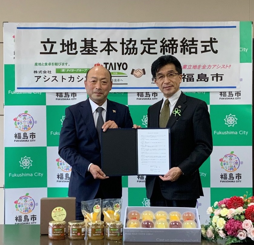 「立地基本協定締結式」と書かれたバックパネルの前で、水海取締役と市長が一緒に協定書を持ち並んでいる写真