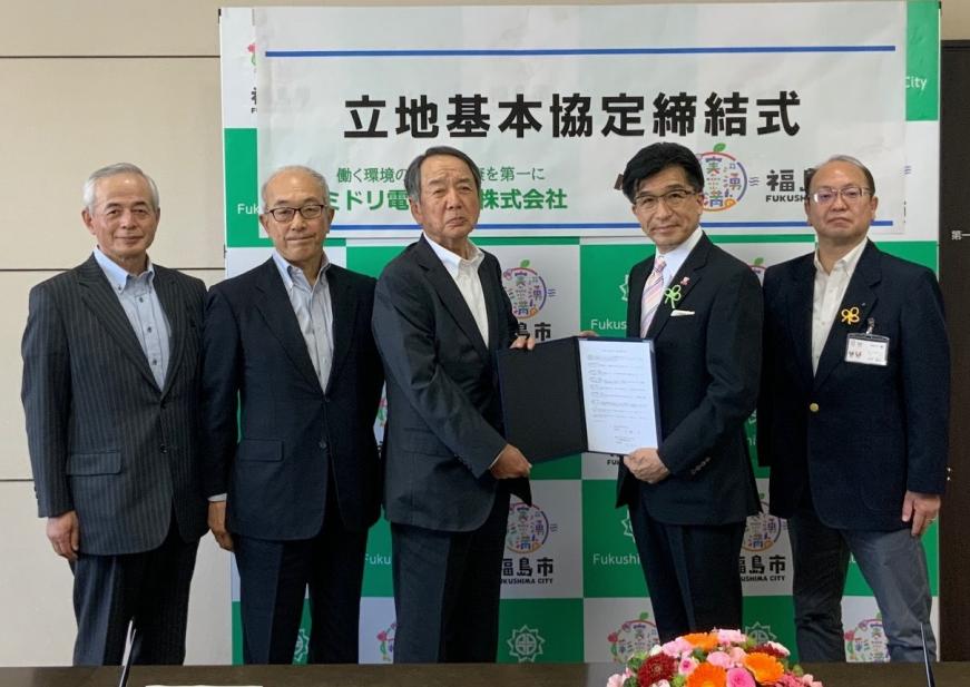 福島市のバックパネルの前で、中央に松村社長と市長が一緒に協定書を持ち立っている両脇に3名の関係者が立っている写真