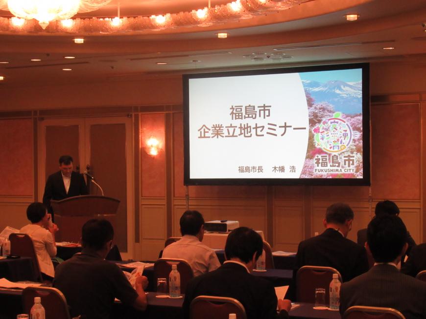 前方に設置されたスクリーンに「福島市企業立地セミナー」と書かれた資料が映し出され、照明を落とした会場の檀上に立つ男性の話を参加者が聞いている様子の写真
