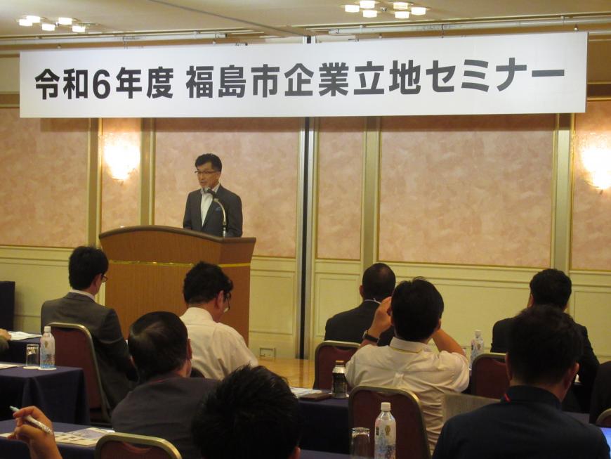 「令和6年度福島市企業立地セミナー」と書かれた横断幕が掲げられた会場の檀上に立つ男性の話を、参加者たちが聞いている様子の写真