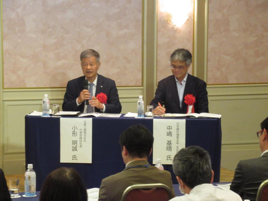左胸に胸章リボンを付けた小形氏と中嶋氏が並んで座り、小形氏がマイクを持って話をしている様子の写真