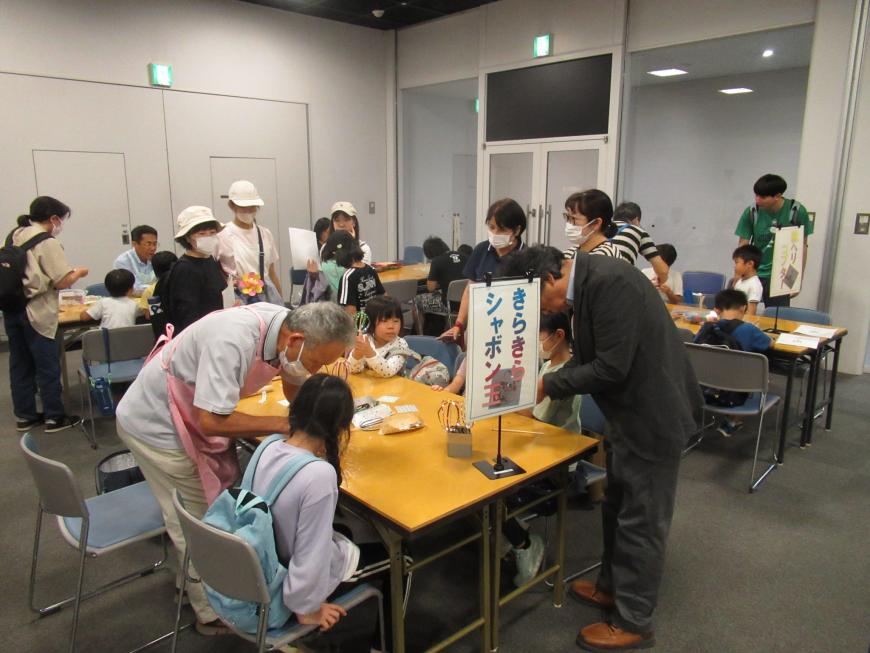子供たちがピンク色のエプロンをつけた男性から指導を受けている「きらきらシャボン玉」のコーナーの写真