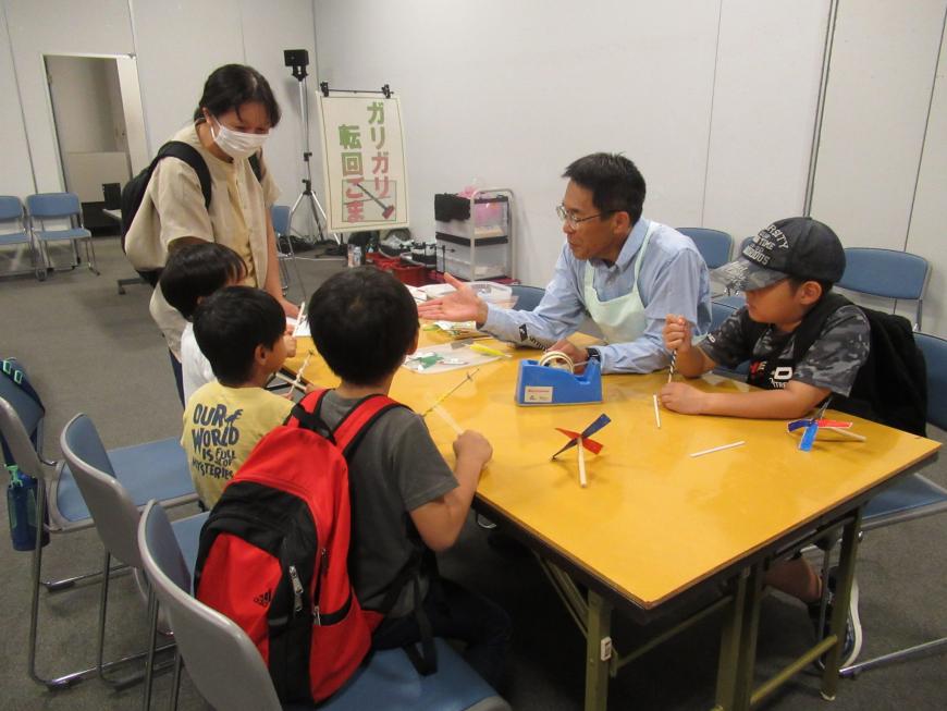 薄い緑色のエプロンを着用した男性から4名の子供たちが作り方を教わっている「ガリガリ回転ごま」のコーナーの写真