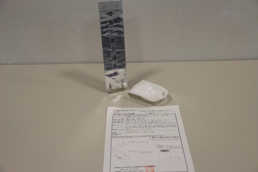 説明書の上に白色のプラスチック製で内側に窪みのある「形が変わる氷入れスコップ」と題した、議長賞を受賞した小学校の部の作品の写真