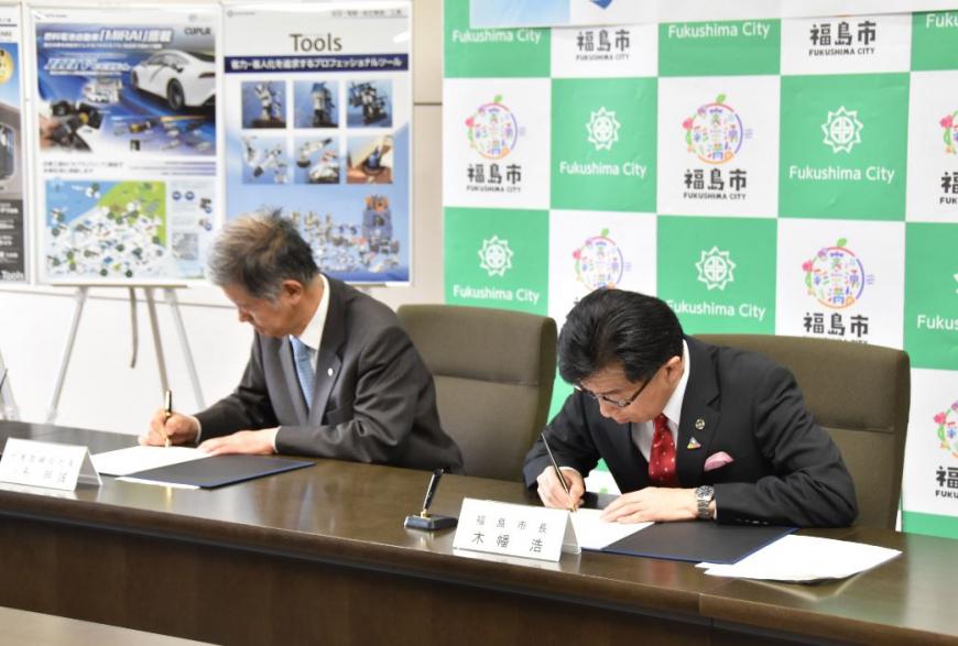 福島市のバックパネルの前に設置された机に並んで座っている小形明誠社長と市長が、協定書に署名を行っている様子の写真