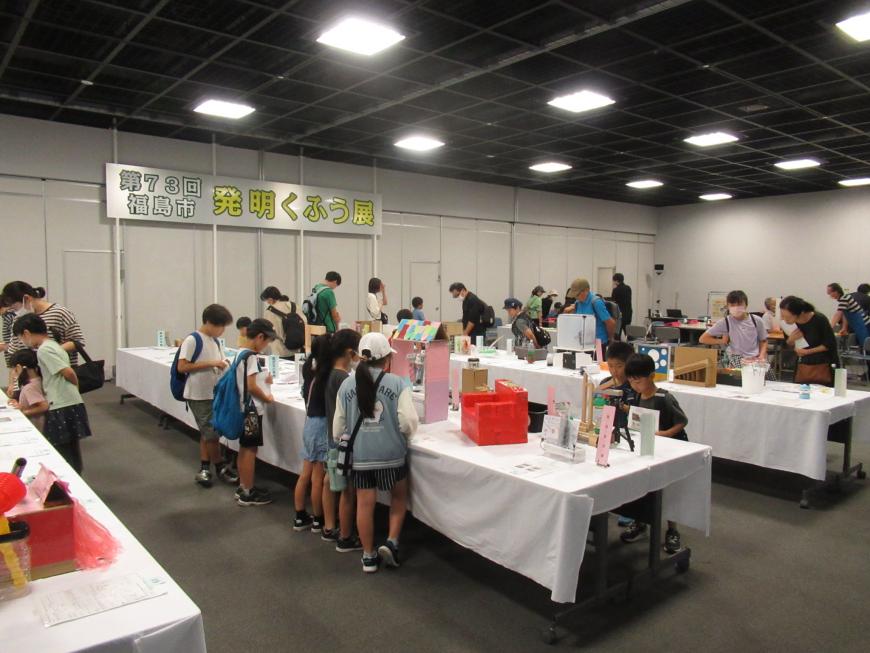 「第73回福島市発明くふう展」と書かれた会場に設置された白いクロスが掛けられた机の上に沢山の作品が展示され、親子連れや子供たちが熱心に見て回っている様子の写真