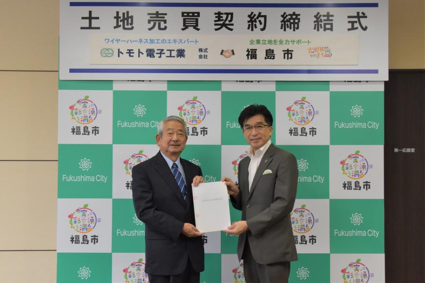 土地売買契約締結式と書かれたバックパネルの前で、滝田社長と市長が一緒に協定書を持ち立っている写真
