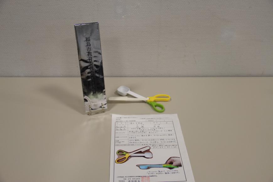 説明書の上に緑と黄色の持ち手でハサミの様な形をした先にスプーンとヘラが取り付けられた「片手で計量スプーンのすり切りが出来る! 「片手ではかる君」」と題した、教育長賞を受賞した小学校の部の作品の写真