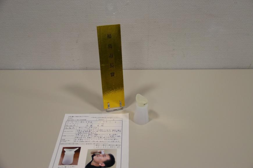 説明書の上に、洗顔液の様な形の下にプラスチック製の土台が作られた「誰でも上手に点眼できる!「アイ・ピタット」」と題した市長賞を受賞した小学校の部の作品の写真