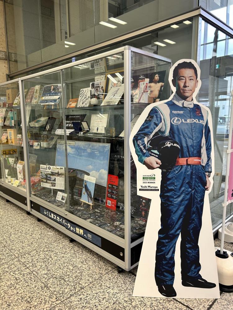 ショーケースの前に飾られたフライトスーツを着てヘルメットを持った室屋選手の等身大パネルの写真