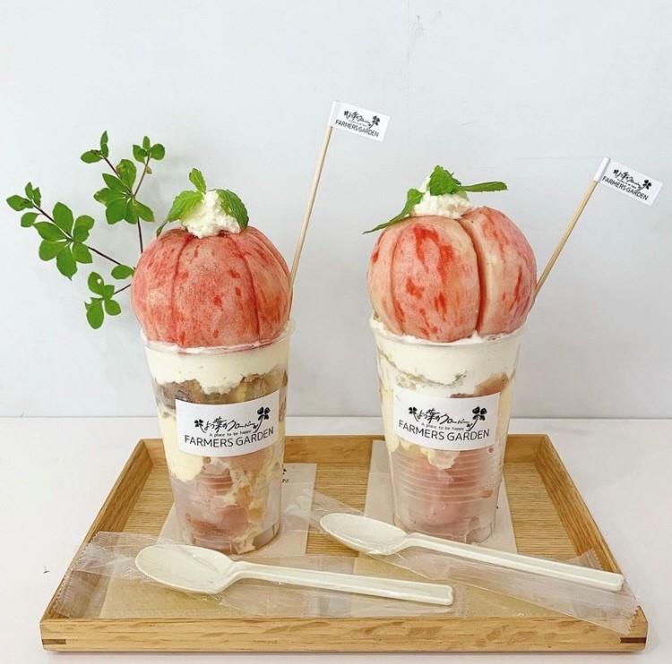 木製トレイの上に店の名前が書かれたラベルが貼られた透明の容器の上に桃がまるごと一個のっている2つのパフェの写真