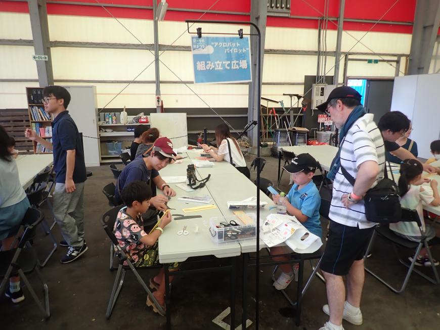 「組み立て広場」と書かれた体験ブースで、親に見守られながら子供たちがアクロバキット飛行機を作っている様子の写真