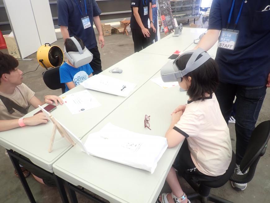 長机の椅子に向かい合って座ったVRゴーグルをつけた男の子と女の子が、VR飛行の体験を行っている様子の写真